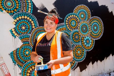 Aboriginal woman holding paint pot in front of colourful Aboriginal mural